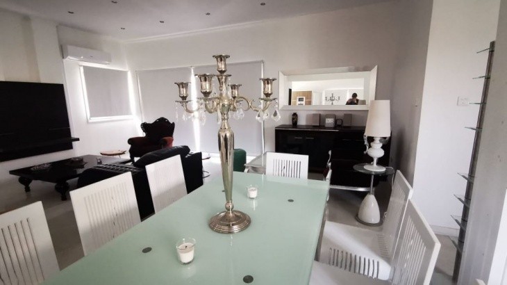 the space of the living room, with the dining table with a big candlestick on top, white chairs, a flat tv, and two sofa chairs
